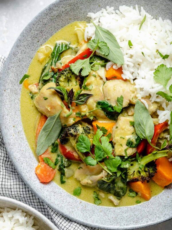 Thai green curry chicken in a bowl with fluffy white rice, topped with fresh cilantro & Thai basil. The bowl sit atop a light blue surface, next to a checkered blue & white linen napkin & a second bowl of green curry.