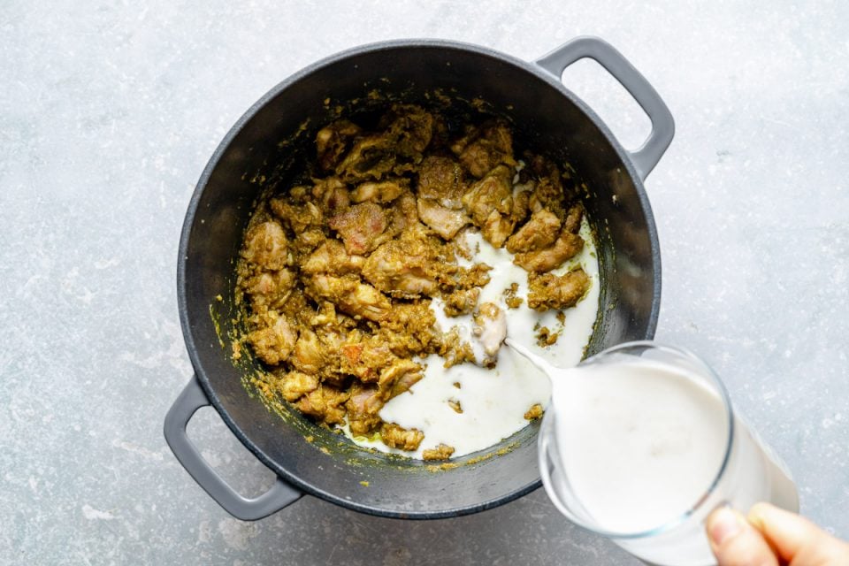 How to make Thai green curry, Step 6: Pouring coconut milk over green curry chicken in a large Dutch oven. The Dutch oven sits atop a light blue surface.