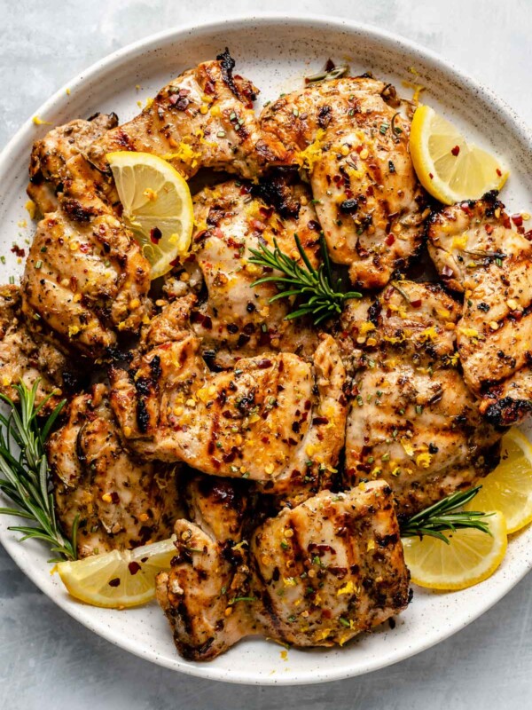 Grilled Tuscan chicken thighs shown on a white speckled plate atop a light blue surface. The chicken is garnished with lemon wedges, fresh rosemary, & crushed red pepper flake.