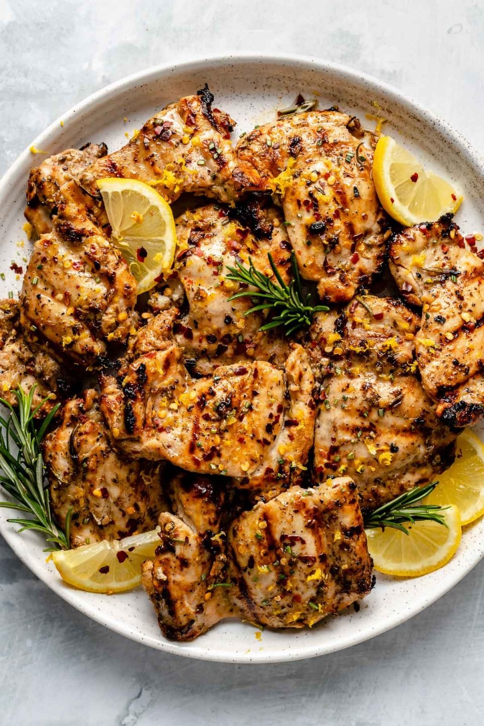 Grilled Tuscan chicken thighs shown on a white speckled plate atop a light blue surface. The chicken is garnished with lemon wedges, fresh rosemary, & crushed red pepper flake.