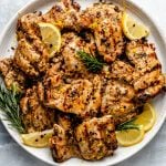 Grilled Tuscan chicken thighs shown on a white speckled plate atop a light blue surface. The chicken is garnished with lemon wedges, fresh rosemary, & crushed red pepper flake.