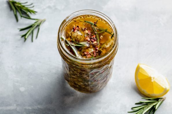 Tuscan marinade shown in a small ball jar, sitting atop a light blue surface with a lemon wedge in the foreground & fresh rosemary sprigs in the background.