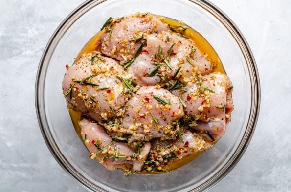 Chicken thighs in a large glass mixing bowl, marinating in Tuscan marinade. The bowl sits atop a light blue surface.