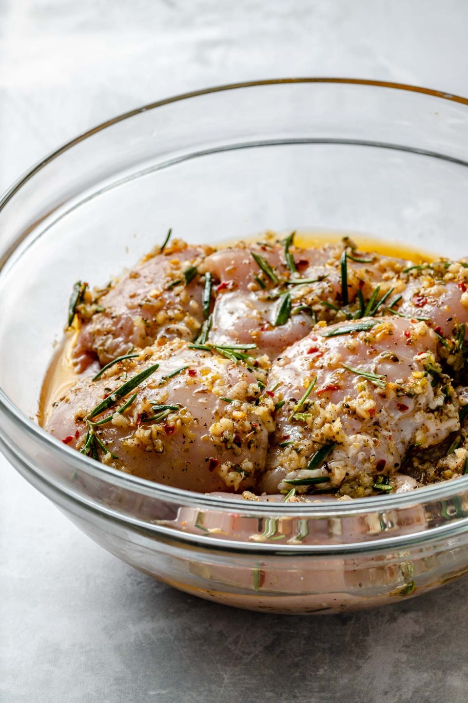 Side angle of chicken thighs in a large glass mixing bowl, marinating in Tuscan marinade. The bowl sits atop a light blue surface.