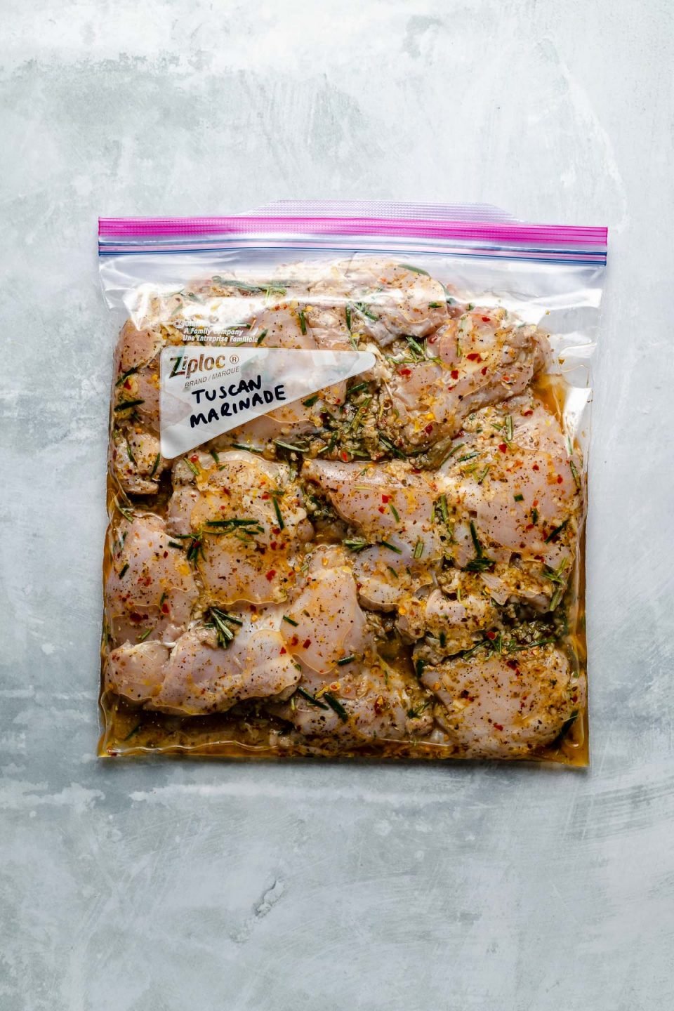 Chicken thighs in a Ziploc bag, marinating in Tuscan marinade. The bag sits atop a light blue surface. "Tuscan Marinade" is penned in the bag's memo area.