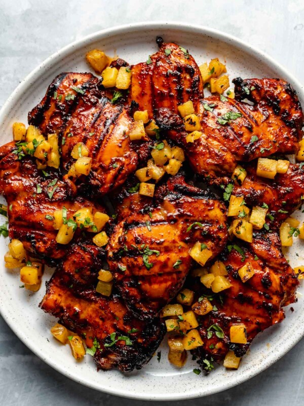 Grilled al Pastor chicken thighs shown on a white speckled plate atop a light blue surface. The chicken is garnished with finely chopped herbs & grilled pineapple.