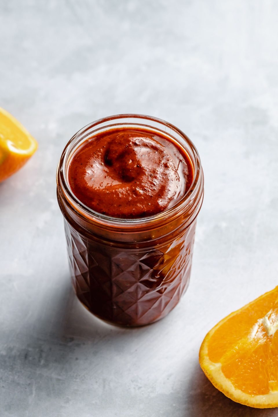 Al Pastor marinade shown in a small ball jar, sitting atop a light blue surface with an orange slice resting on the surface in the foreground & the background.