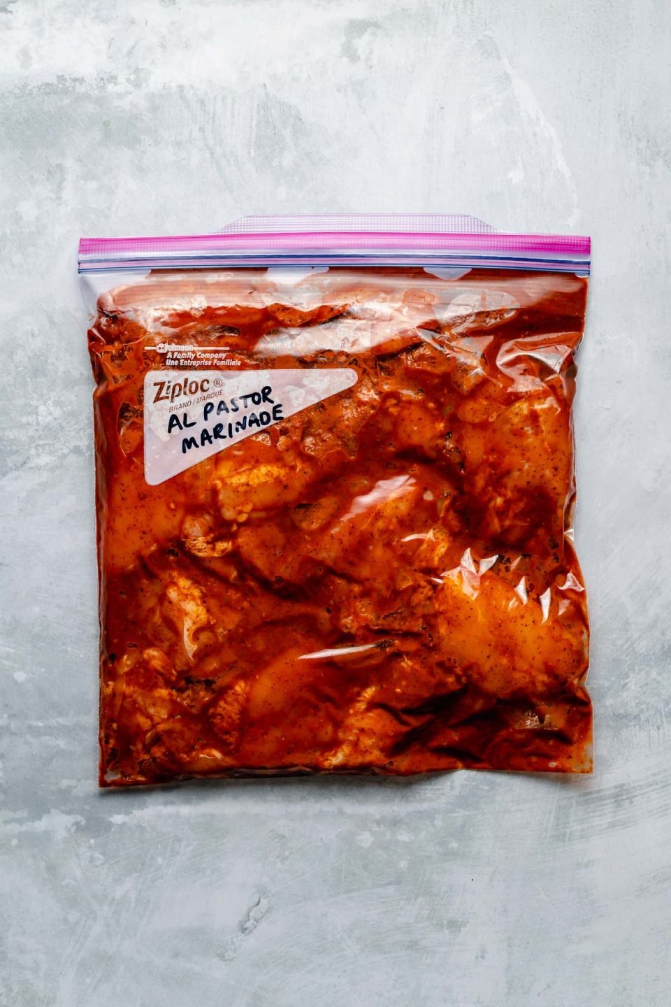 Chicken thighs in a Ziploc bag, marinating in al Pastor marinade. The bag sits atop a light blue surface. “Al Pastor Marinade" is penned in the bag's memo area.
