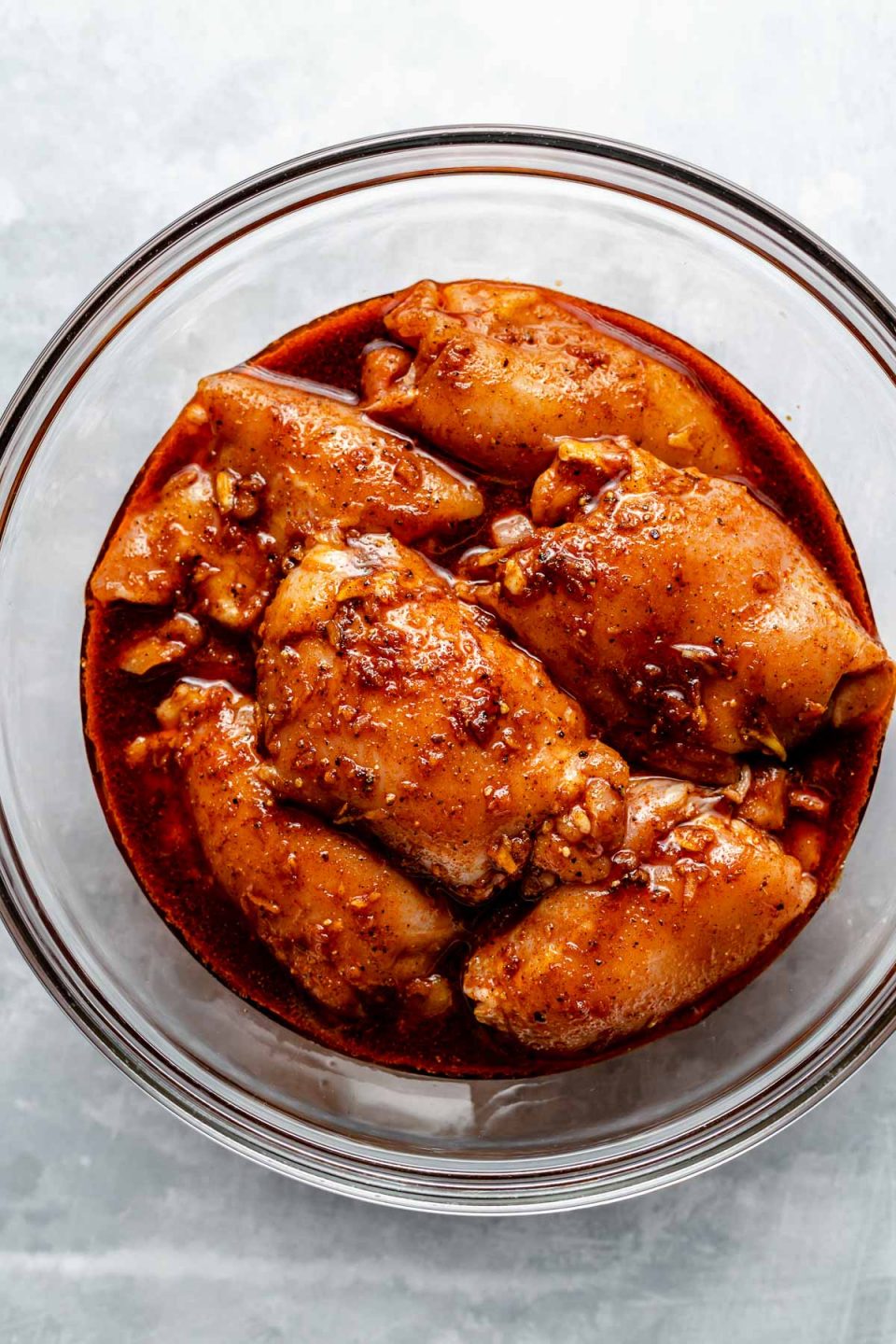 Chicken thighs in a large glass mixing bowl, marinating in All-Purpose marinade. The bowl sits atop a light blue surface.