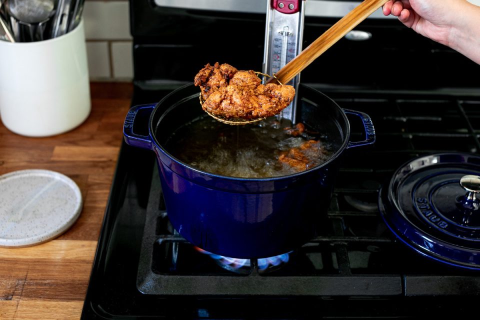 An angled shot of crispy, golden brown fried chicken being pulled out of a blue Staub Tall Cocotte, with a spider strainer, filled with herb & garlic infused frying oil. The blue cocotte sits on top of a gas stovetop range and the lid to the cocotte sits on the range beside it.
