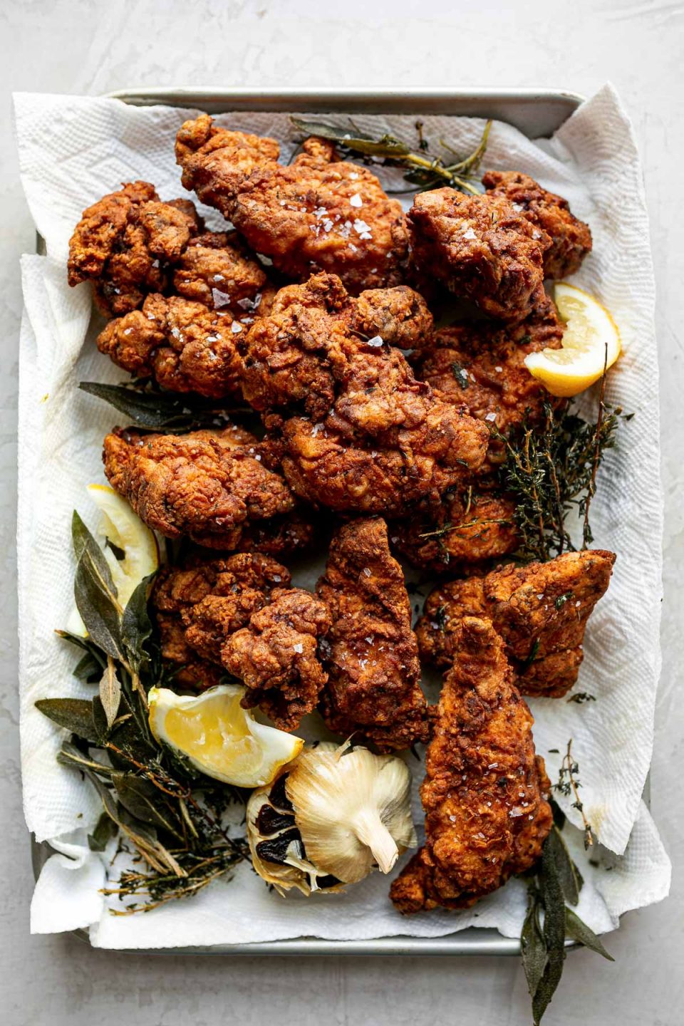 A top down shot of pieces of crispy, golden brown fried chicken, sprinkled in flaky salt, piled onto a quarter sheet pan lined with paper towels. Leftover herbs & garlic from the frying oil and lemon slices resting next to and around the pieces of chicken.