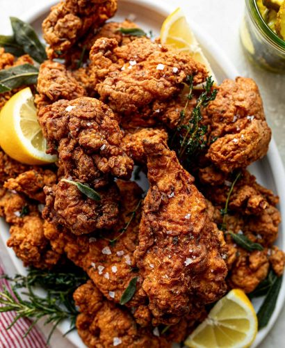 A top down shot of crispy, golden brown fried chicken is piled high on a white oval platter. Sprigs of herbs and slices of lemon are intertwined with the chicken. The platter sits on a white textured surface with a red & white striped linen napkin. A jar of pickles sits to the right of the platter.