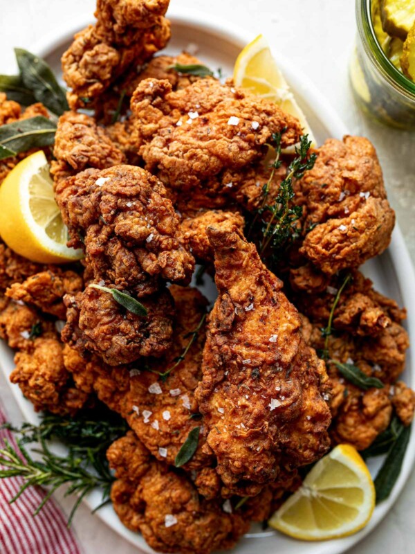 A top down shot of crispy, golden brown fried chicken is piled high on a white oval platter. Sprigs of herbs and slices of lemon are intertwined with the chicken. The platter sits on a white textured surface with a red & white striped linen napkin. A jar of pickles sits to the right of the platter.