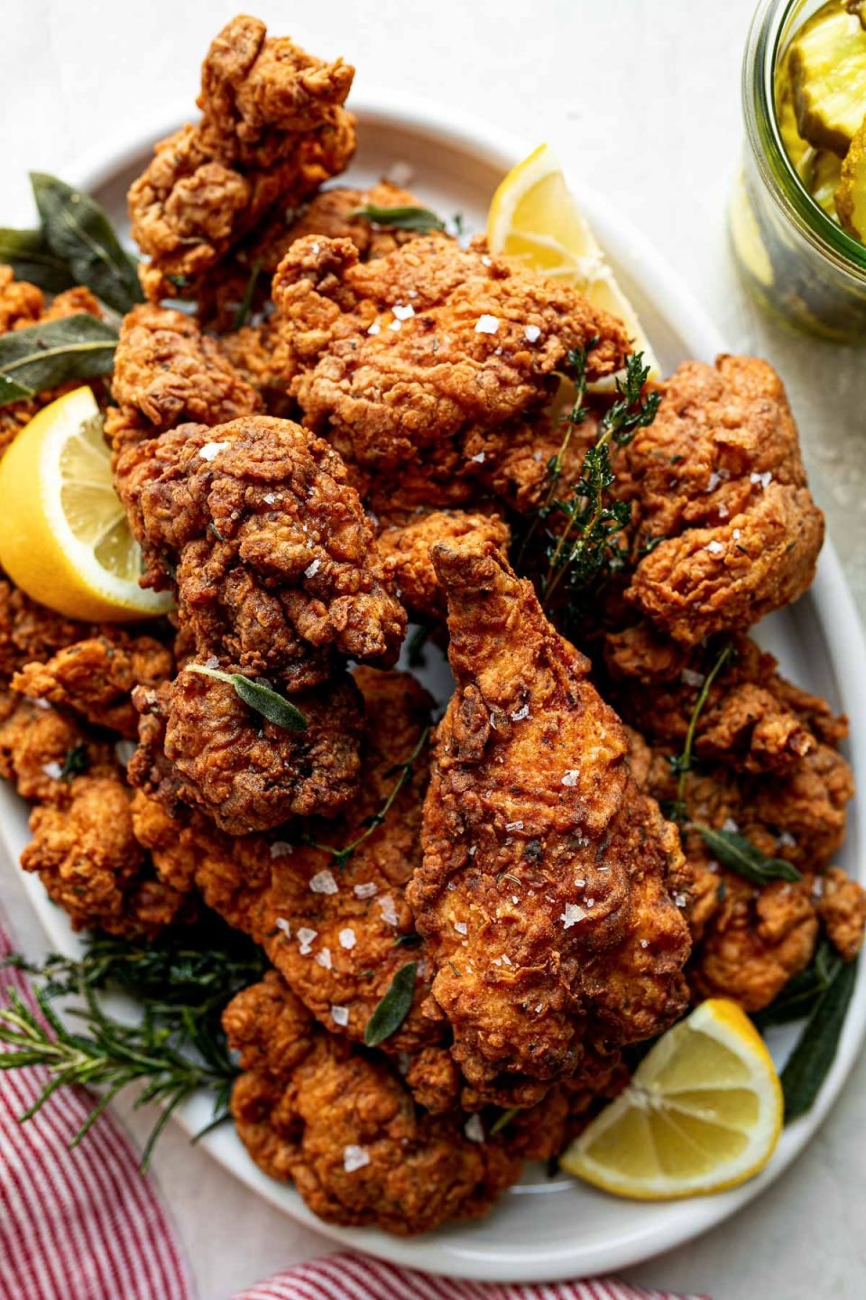 A top down shot of crispy, golden brown fried chicken is piled high on a white oval platter. Sprigs of herbs and slices of lemon are intertwined with the chicken. The platter sits on a white textured surface with a red & white striped linen napkin. A jar of pickles sits to the right of the platter.