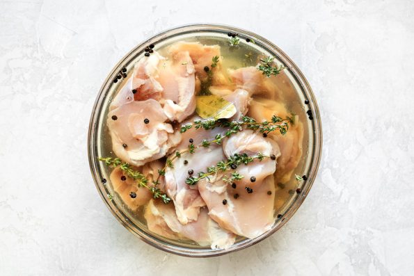 Boneless, skinless chicken thighs brining in a simple poultry brine in a clear glass mixing bowl atop a creamy white surface.