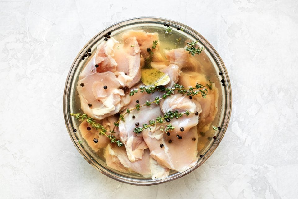 A top down shot of a bowl filled with raw chicken in a simple brine filled with peppercorn & fresh poultry herbs.