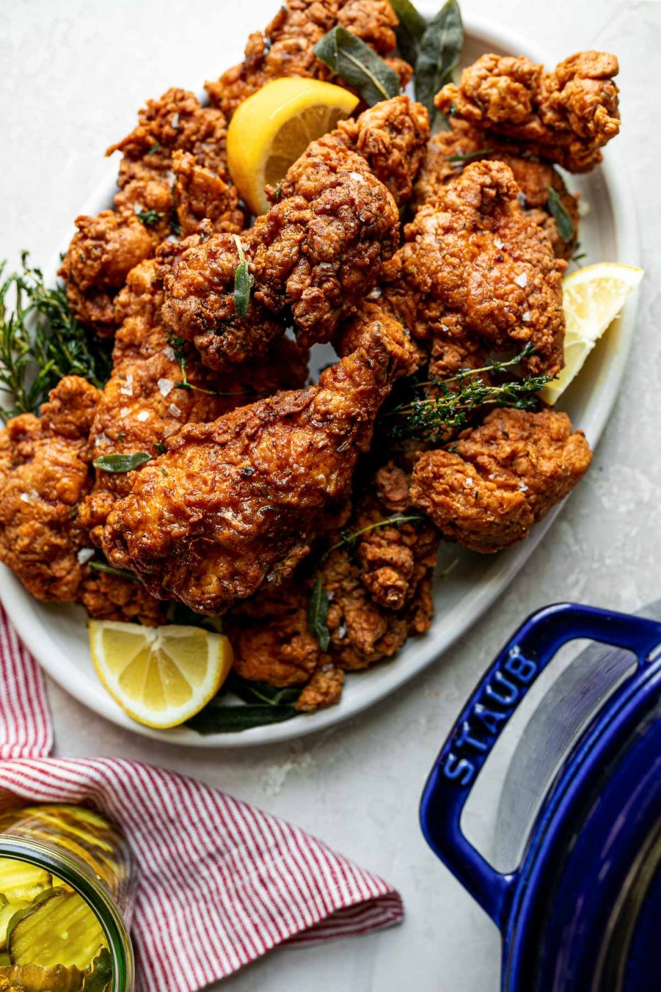 A top down shot of crispy, golden brown fried chicken is piled high on a white oval platter. Sprigs of herbs and slices of lemon are intertwined with the chicken. The platter sits on a white textured surface with a red & white striped linen napkin, a jar of pickles, and a blue Staub tall cocotte rests in front of the platter.