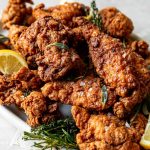 An angled shot of crispy, golden brown fried chicken is piled high on a white oval platter. Sprigs of herbs and slices of lemon are intertwined with the chicken. The platter sits on a white textured surface with a red & white striped linen napkin is tucked in front of the platter.