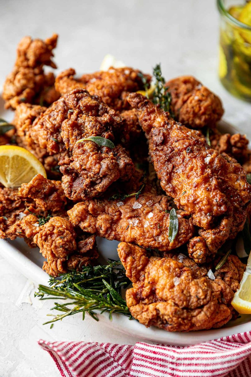 An angled shot of crispy, golden brown fried chicken is piled high on a white oval platter. Sprigs of herbs and slices of lemon are intertwined with the chicken. The platter sits on a white textured surface with a red & white striped linen napkin is tucked in front of the platter and a jar of pickles sit behind the platter.
