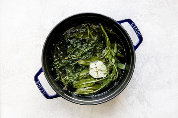 A top down shot of a blue Staub Tall Cocotte filled with hot herb & garlic infused frying oil sits on top of a white textured surface.