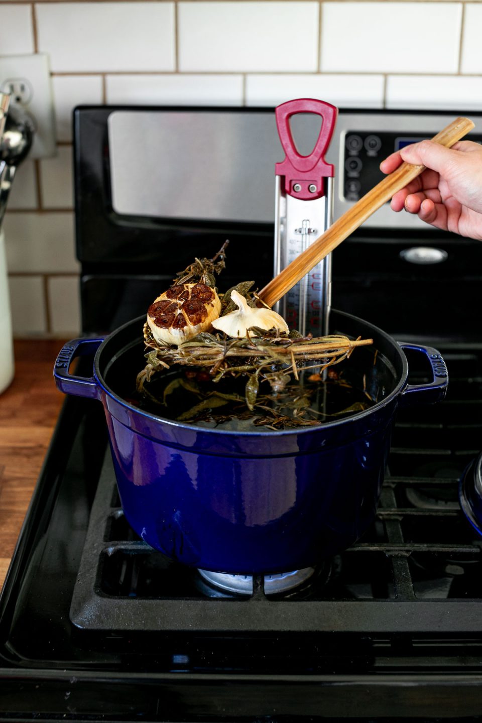 An angled shot of a piece of browned herbs & garlic being pulled out of a blue Staub Tall Cocotte, with a spider strainer, filled with herb & garlic infused frying oil. A candy thermometer rests inside the cocotte. The blue cocotte sits on top of a gas stovetop range.