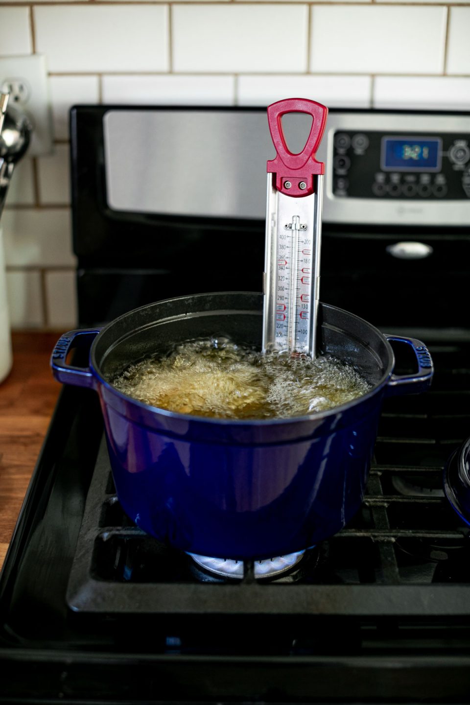 An angled shot of a single blue Staub Tall Cocotte sits on top of a gas stovetop range filled with boiling herb & garlic infused frying oil. Inside the oil pieces of fried chicken are being deep fried. A candy thermometer rests inside the cocotte to watch the temperature of the frying oil.
