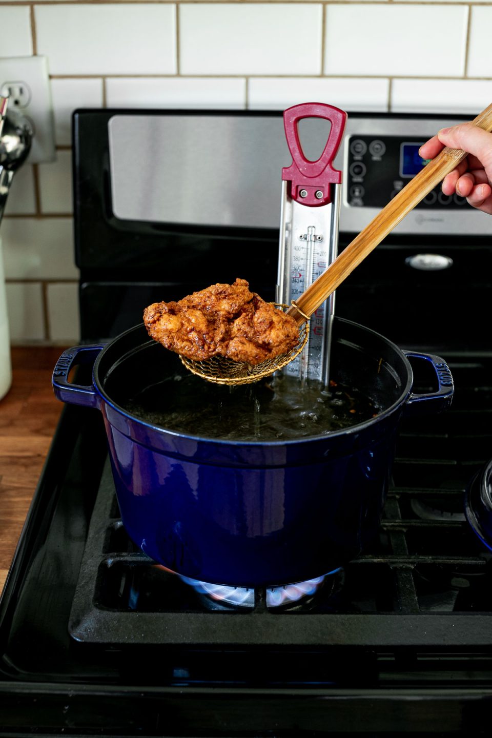 An angled shot of a piece of crispy, golden brown fried chicken being pulled out of a blue Staub Tall Cocotte, with a spider strainer, filled with herb & garlic infused frying oil. A candy thermometer rests inside the cocotte. The blue cocotte sits on top of a gas stovetop range and the lid to the cocotte sits on the range beside it.