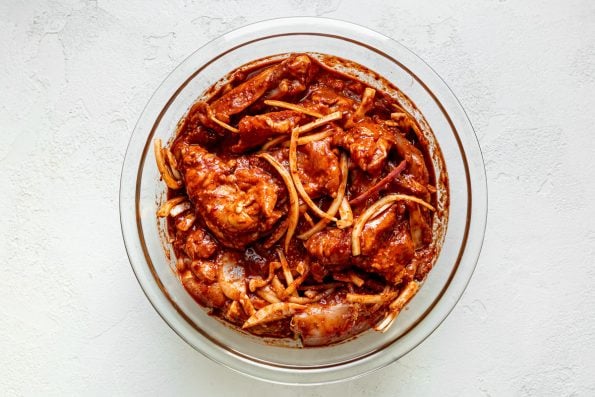 Chicken thighs and thinly sliced onion marinate in al pastor marinade in a large glass mixing bowl atop a white plaster surface.