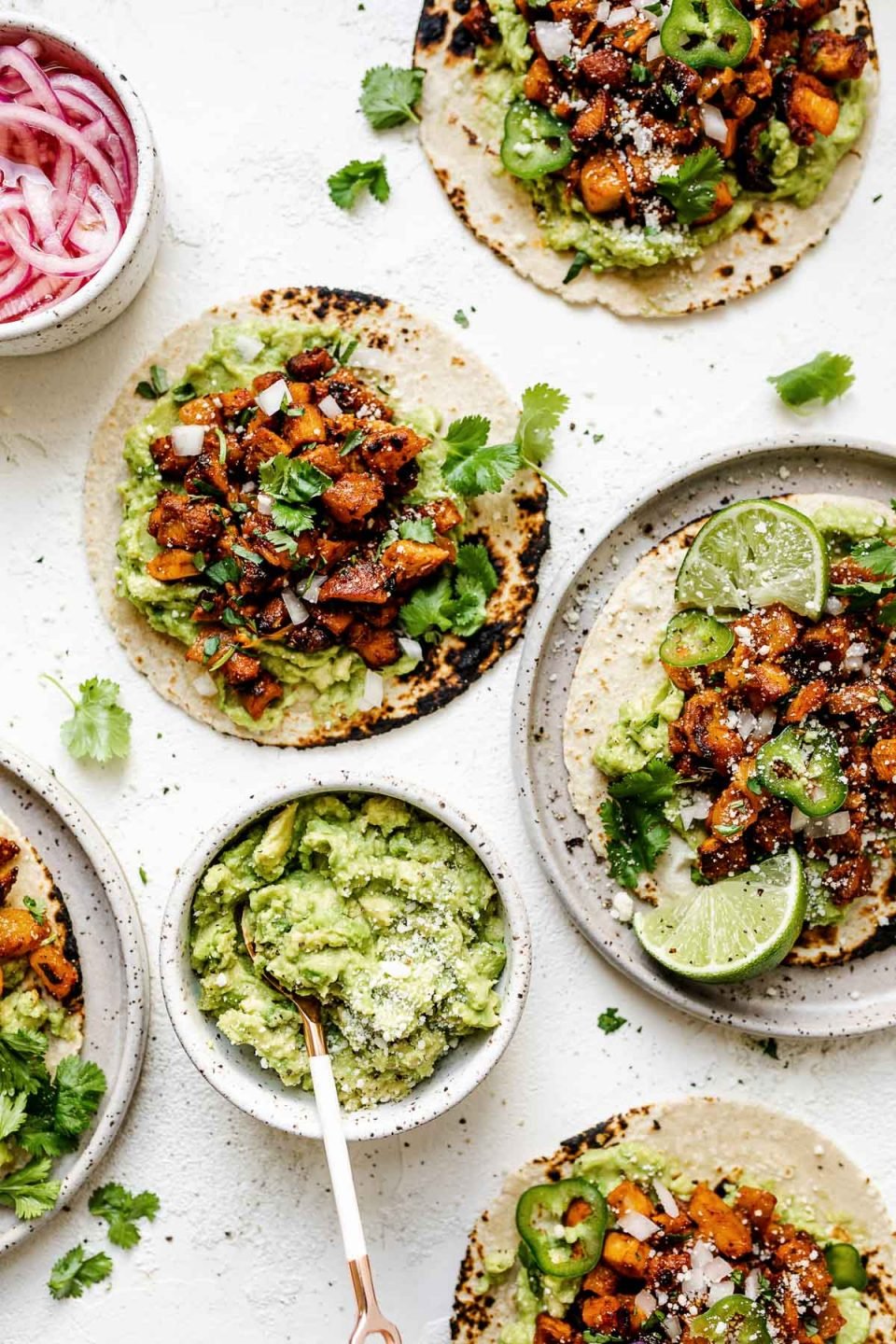 Five chicken al pastor tacos on a white plaster surface. Two of the tacos are placed on small speckled ceramic plates. Surrounding the tacos are pickled red onions, mashed avocado, and fresh cilantro leaves.