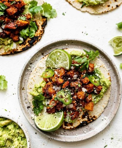 A chicken al pastor taco on a speckled ceramic plate, topped with thinly sliced jalapeno & cotija cheese. The taco is surrounded by 3 other al pastor tacos, a small bowl of mashed avocado, fresh cilantro leaves & lime wedges.