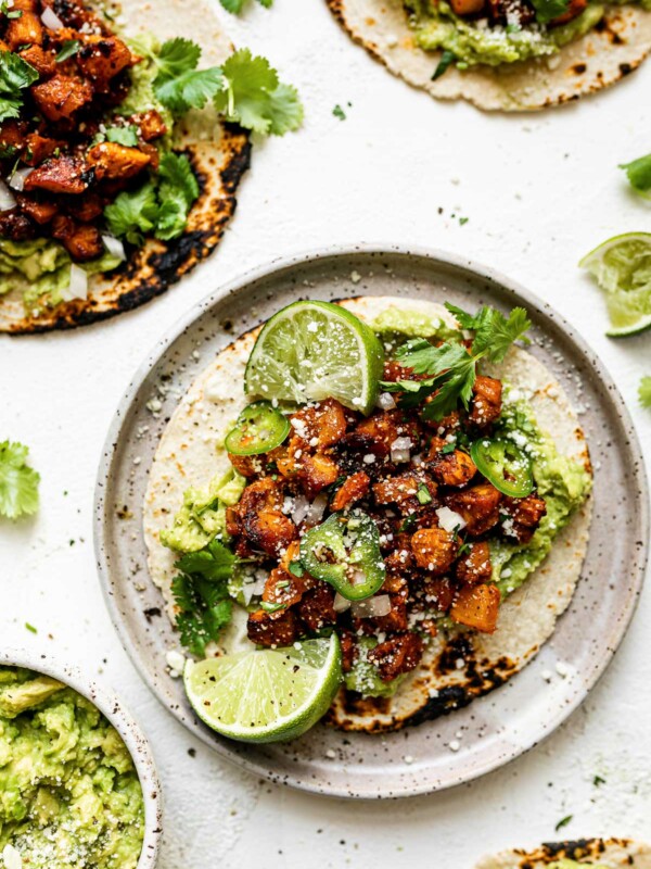 A chicken al pastor taco on a speckled ceramic plate, topped with thinly sliced jalapeno & cotija cheese. The taco is surrounded by 3 other al pastor tacos, a small bowl of mashed avocado, fresh cilantro leaves & lime wedges.
