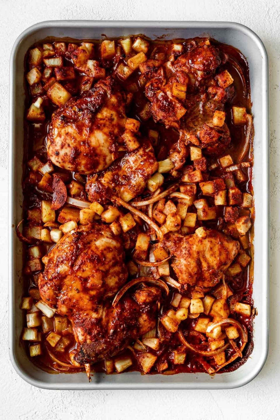 Baked al pastor chicken thighs & sliced onion on a silver quarter baking sheet with finely diced pineapple. The sheet pan sits atop a white plaster surface.