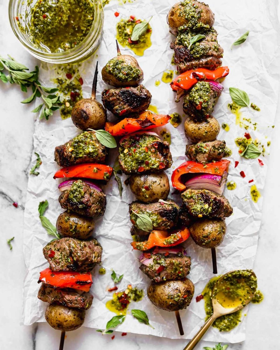 Three Chimichurri Steak & Potato Kebabs lie flat on a light gray surface surrounded by fresh herbs, an open jar of chimichurri sauce & a gold spoon resting that had been used to drizzle chimichurri sauce on the kebabs