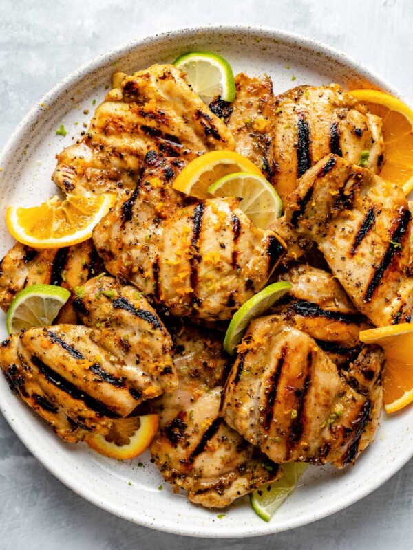 Grilled mojo chicken thighs on a speckled white plate atop a light blue surface. The chicken thighs are garnished with orange & lime wedges & zest.