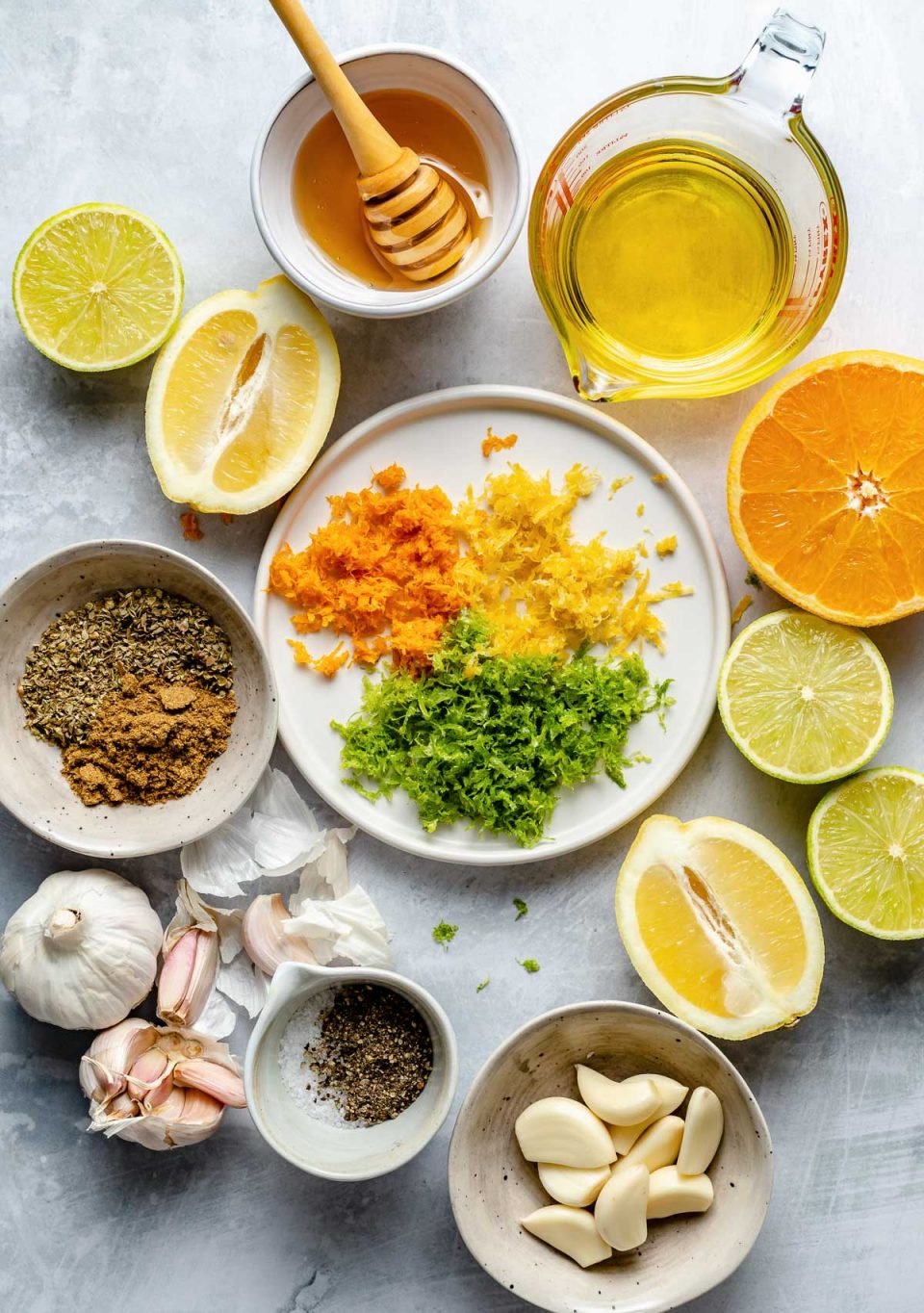 Mojo marinade ingredients arranged on a light blue surface: oranges, lemons, limes, zest of all 3, garlic, cumin, salt, pepper, honey, & olive oil.