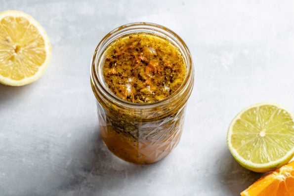Mojo marinade shown in a small jar atop a light blue surface. A halved lime & orange wedge in the foreground, & a halved lemon in the background.