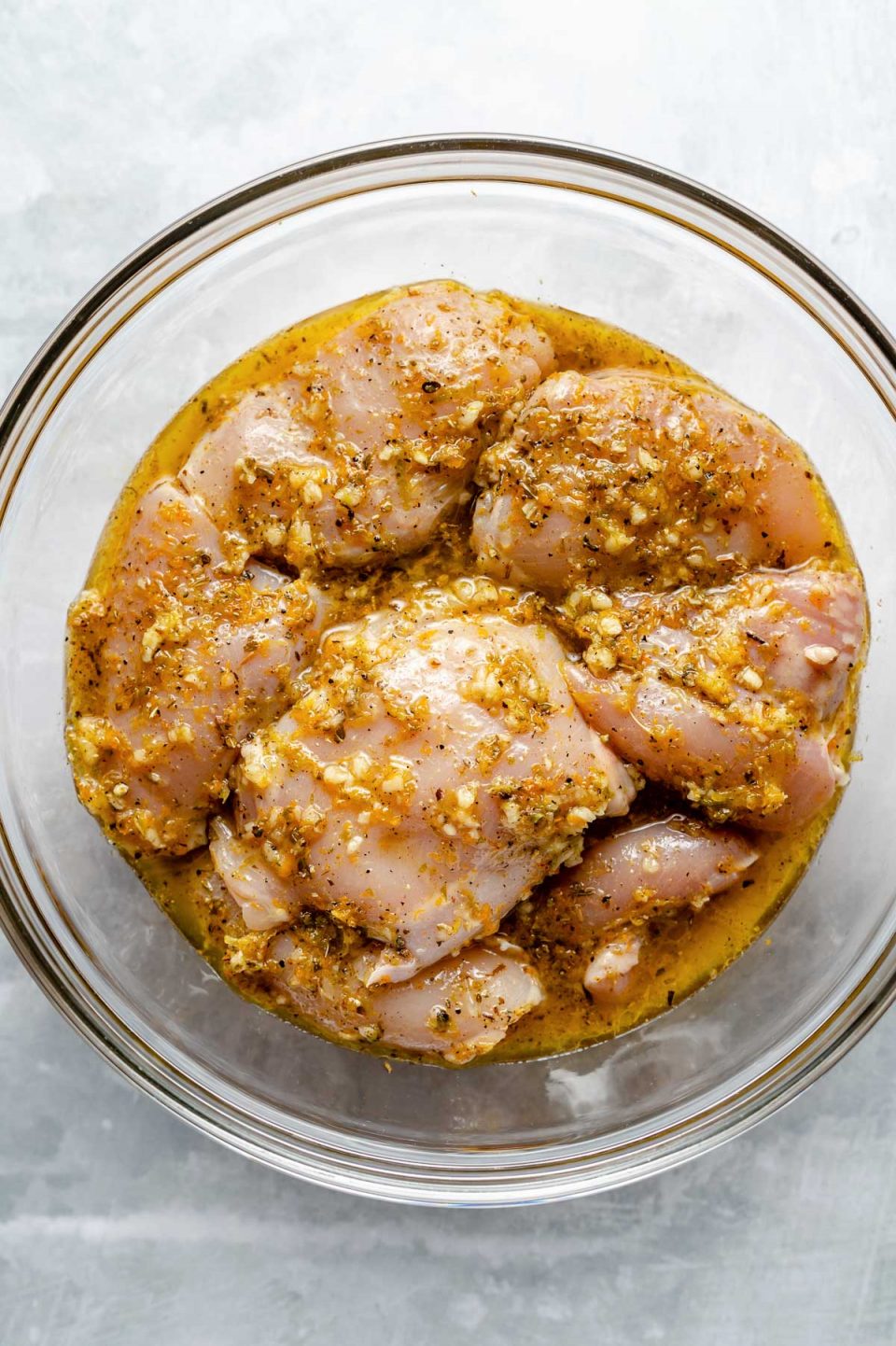 Chicken thighs in mojo marinade in a large glass bowl atop a light blue surface.