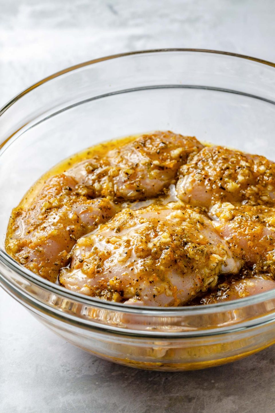 Side angle of chicken thighs in mojo marinade in a large glass bowl atop a light blue surface.