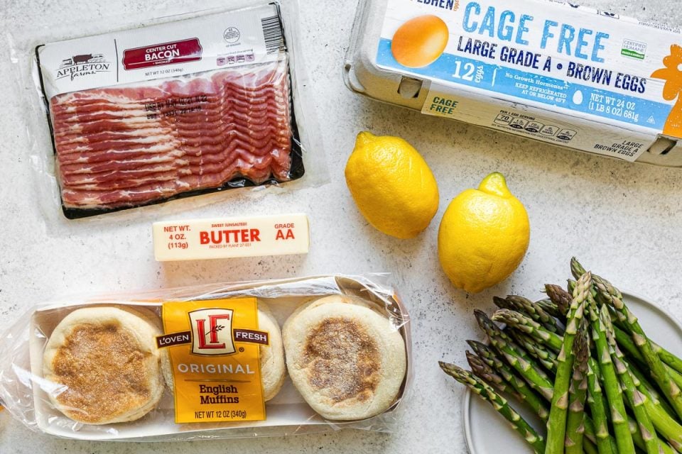 Eggs benedict ingredients from ALDI - Appleton Farms Center-Cut Bacon, L'Oven Fresh English Muffins, lemons, butter, asparagus & Gold Hen Cage-Free Brown Eggs - atop a white surface.