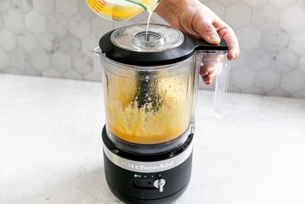 Blender hollandaise sauce - a woman's hands operating & slowly streaming butter into a blender to make easy hollandaise sauce. The blender sits atop a white surface in front of a tiled surface.