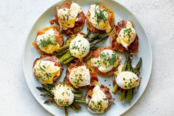 Eggs benedict variations with asparagus, bacon, prosciutto & smoked salmon atop a light blue speckled platter, atop a white surface.