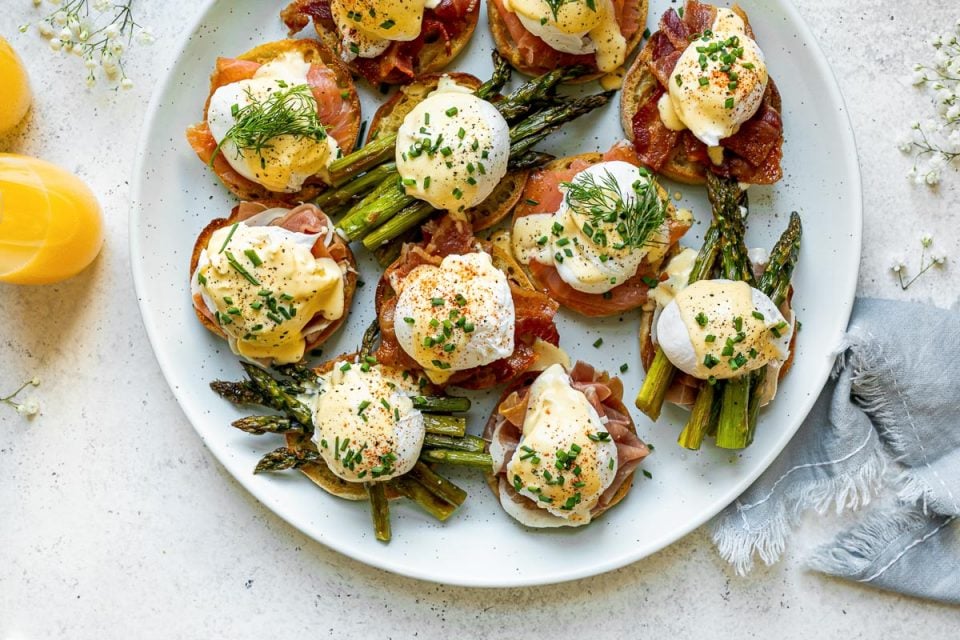 Eggs benedict variations with asparagus, bacon, prosciutto & smoked salmon atop a light blue speckled platter, atop a white surface. Next to the platter are baby's breath flowers, champange flutes with orange juice & a blue linen napkin.
