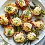 Eggs benedict variations with asparagus, bacon, prosciutto & smoked salmon atop a light blue speckled platter, atop a white surface. Next to the platter are baby's breath flowers & a blue linen napkin.