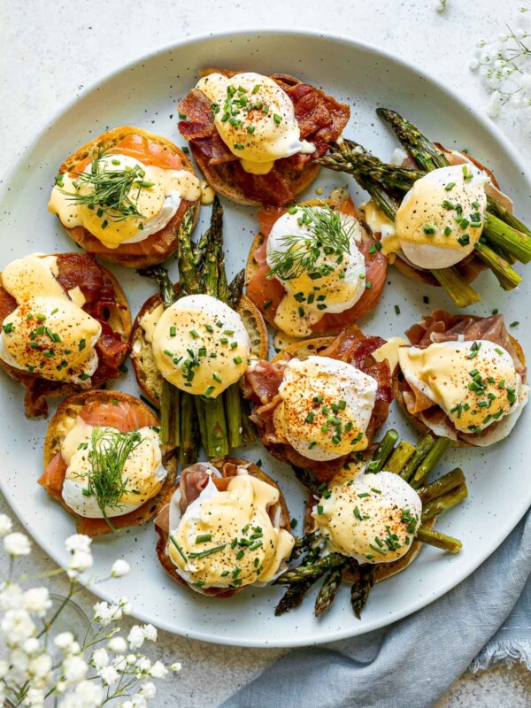 Eggs benedict variations with asparagus, bacon, prosciutto & smoked salmon atop a light blue speckled platter, atop a white surface. Next to the platter are baby's breath flowers & a blue linen napkin.