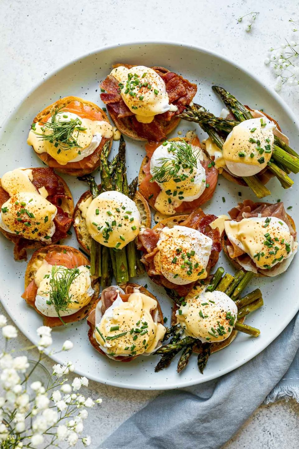 Eggs benedict variations with asparagus, bacon, prosciutto & smoked salmon atop a light blue speckled platter, atop a white surface. Next to the platter are baby's breath flowers & a blue linen napkin.
