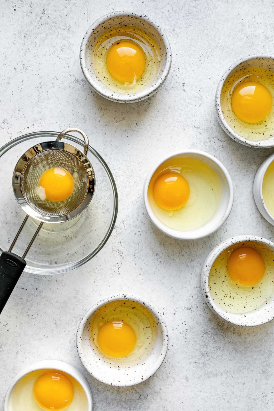 Poached eggs prep: eggs cracked into individual bowls. One egg is cracked in a small strainer to strain the whites. All of the bowls sit atop a white surface.