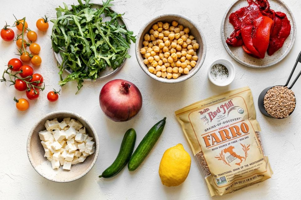 Greek farro salad ingredients arranged on a white surface: tomatoes, feta cheese, arugula, red onion, chickpeas, cucumber, lemon, Bob's Red Mill farro, & roasted red peppers.