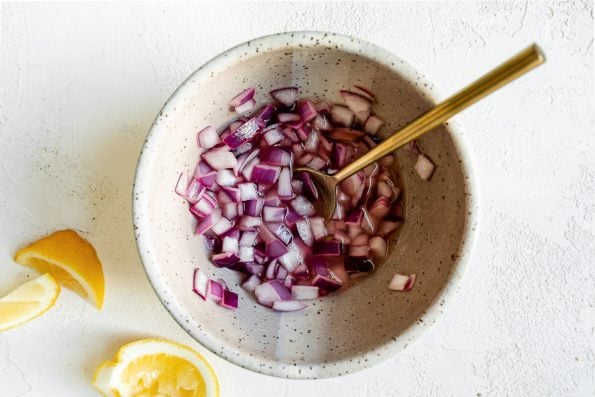 How to make Greek farro salad, Step 2: Diced red onions soaking in lemon juice in a speckled ceramic bowl with a gold spoon. The bowl sits atop a white surface next to a couple of lemon wedges.