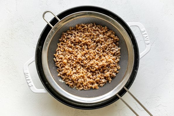 How to make Greek Farro Salad, Step 1: Cook farro. Cooked farro shown in strainer in a white Staub cocotte atop a white surface.