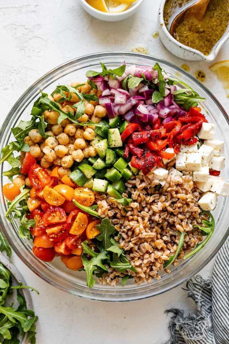 Greek farro salad in large clear glass bowl atop a white surface. The farro salad ingredients are grouped in the bowl - lemony red onions, chickpeas, cucumber, tomatoes, farro, feta, etc. The bowl is surrounded by a small bowl of Greek dressing, a striped gray linen napkin, lemon wedges & a few leaves of baby arugula.